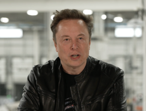 Person in a black leather jacket speaking indoors, industrial background with blurred lights and structures.