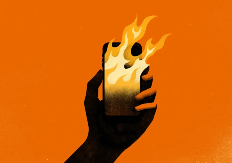 Hand holding a smartphone with flames emerging from the screen against an orange background