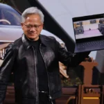 Man in a black leather jacket holding a laptop displaying a digital scene, with a car and robot in the background.