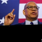 Man in suit gestures while speaking in front of an American flag backdrop.