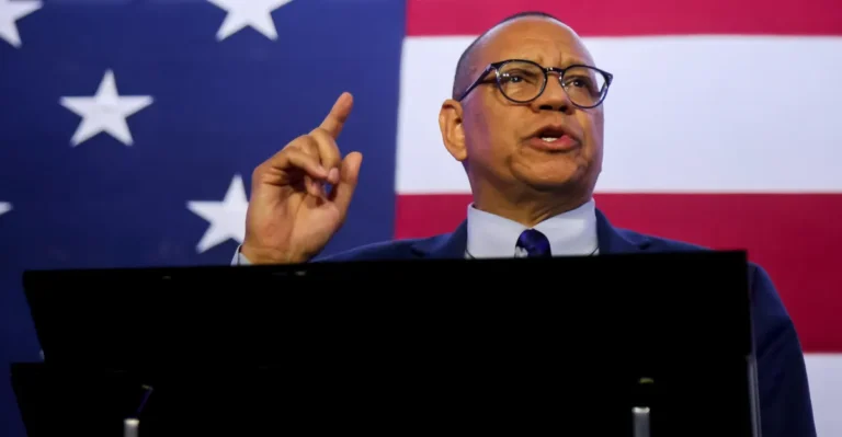 Man in suit gestures while speaking in front of an American flag backdrop.