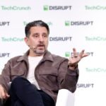 Man speaking with a headset, gesturing with one hand, seated on stage. "TechCrunch Disrupt" text on backdrop.