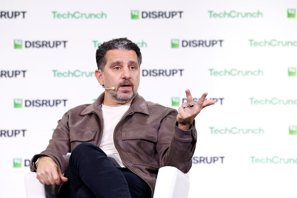 Man speaking with a headset, gesturing with one hand, seated on stage. "TechCrunch Disrupt" text on backdrop.