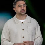 Man in a beige cardigan speaking, blurred green and blue background, appears to be in a presentation setting.