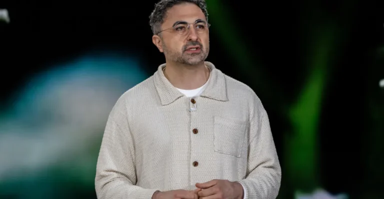 Man in a beige cardigan speaking, blurred green and blue background, appears to be in a presentation setting.