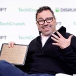 Man with glasses smiles and gestures while holding a microchip on stage, TechCrunch Disrupt logos in the background.