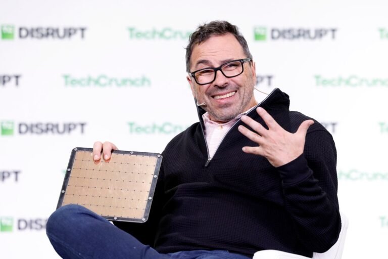 Man with glasses smiles and gestures while holding a microchip on stage, TechCrunch Disrupt logos in the background.