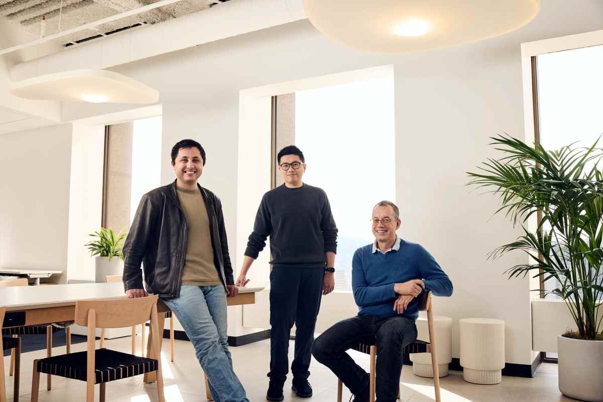 Three men in a modern office setting, two standing and one seated, with tables, chairs, large windows, and potted plants.