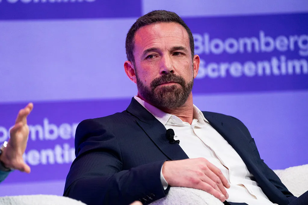 Man in a suit seated on a stage, holding a microphone. Background displays "Bloomberg Screentime.