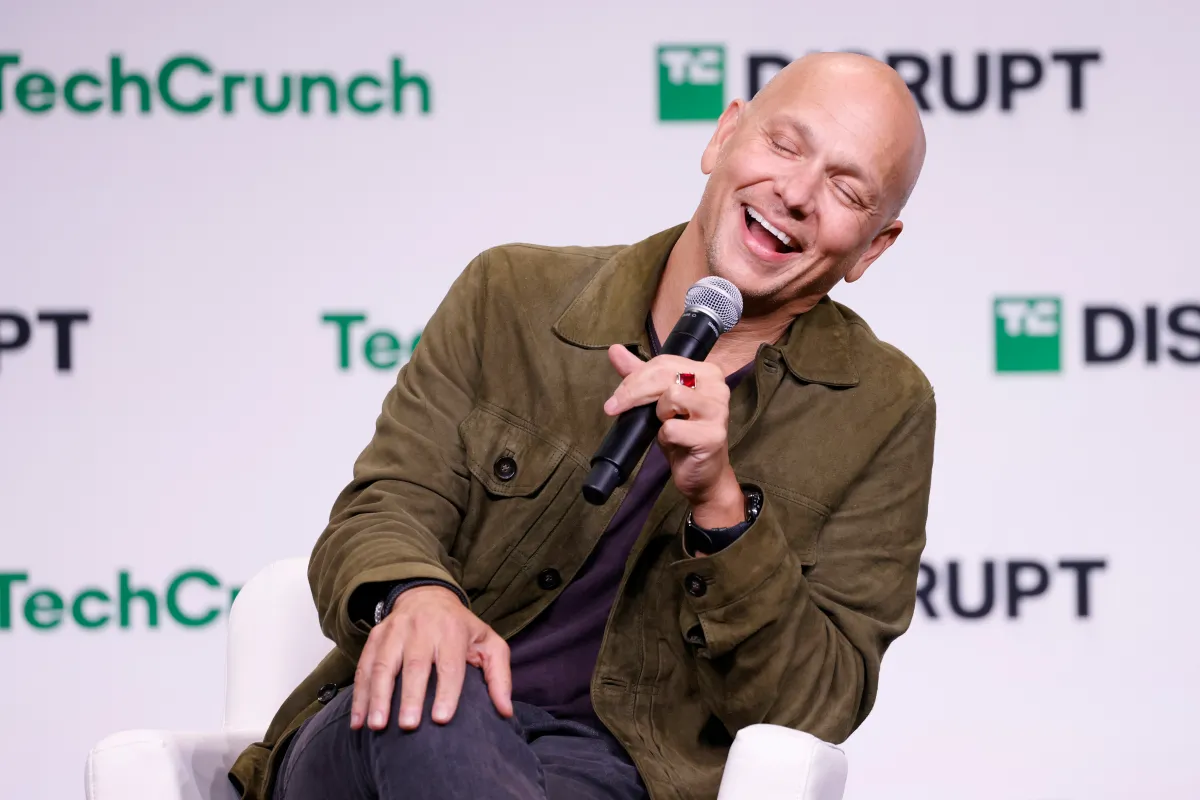 Man in a green jacket holding a microphone, laughing on stage at TechCrunch Disrupt event.