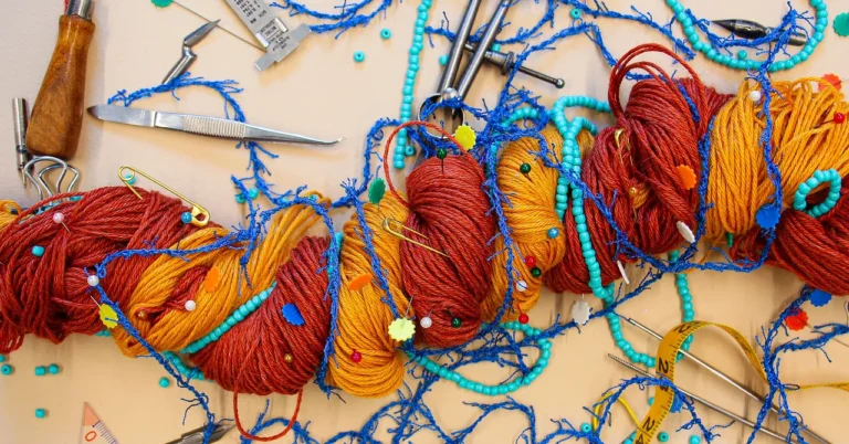Colorful yarn entangled with blue threads, beads, and sewing tools on a beige surface, resembling a complex network.