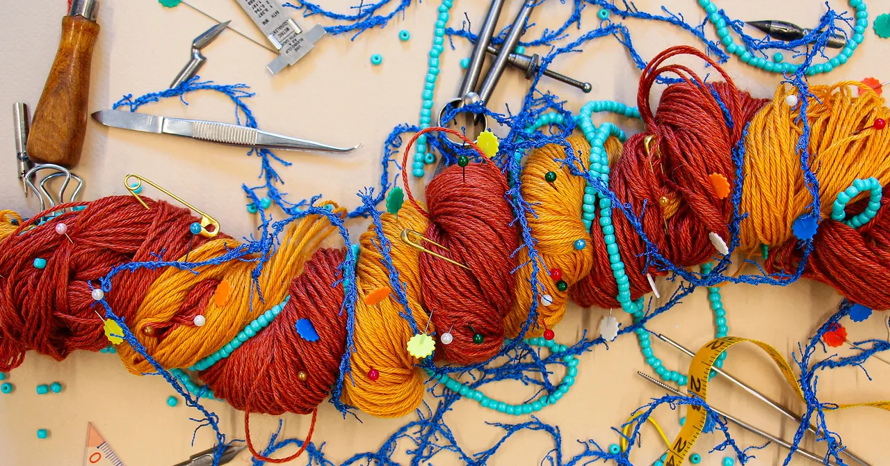 Colorful yarn entangled with blue threads, beads, and sewing tools on a beige surface, resembling a complex network.