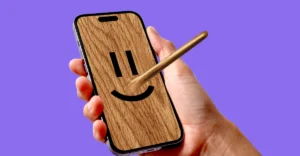 Hand holding a smartphone with a wooden texture, smiling face, and Pinocchio-like nose, against a purple background.