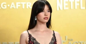 Woman with long dark hair in a lace dress stands against a yellow backdrop with partial Netflix logo visible.