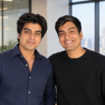 Two men smiling in a modern office with large windows and cityscape background.