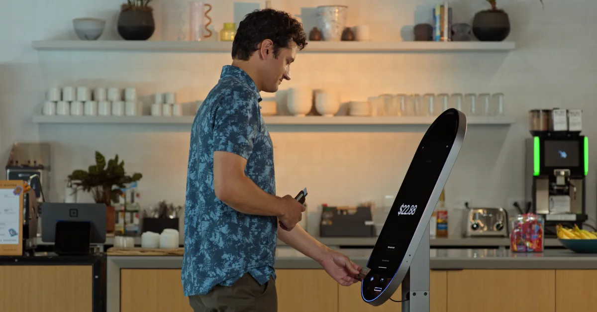 Man using a sleek, curved AI kiosk with a touchscreen displaying $22.88 in a modern cafe setting with shelves and appliances.