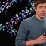 Person speaking on stage with colorful abstract pattern in the background, holding a small device, wearing a blue sweater.