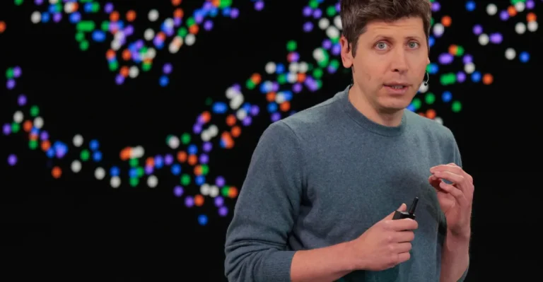 Person speaking on stage with colorful abstract pattern in the background, holding a small device, wearing a blue sweater.