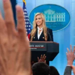 Woman at podium labeled "White House Washington" with raised hands in foreground, US flag and White House emblem behind.