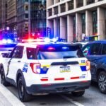 Police cars with flashing lights on a city street lined with tall buildings, parked cars nearby.