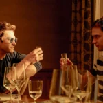 Two men seated at a table, raising glasses, surrounded by wine glasses and floral decorations in a warmly lit room.