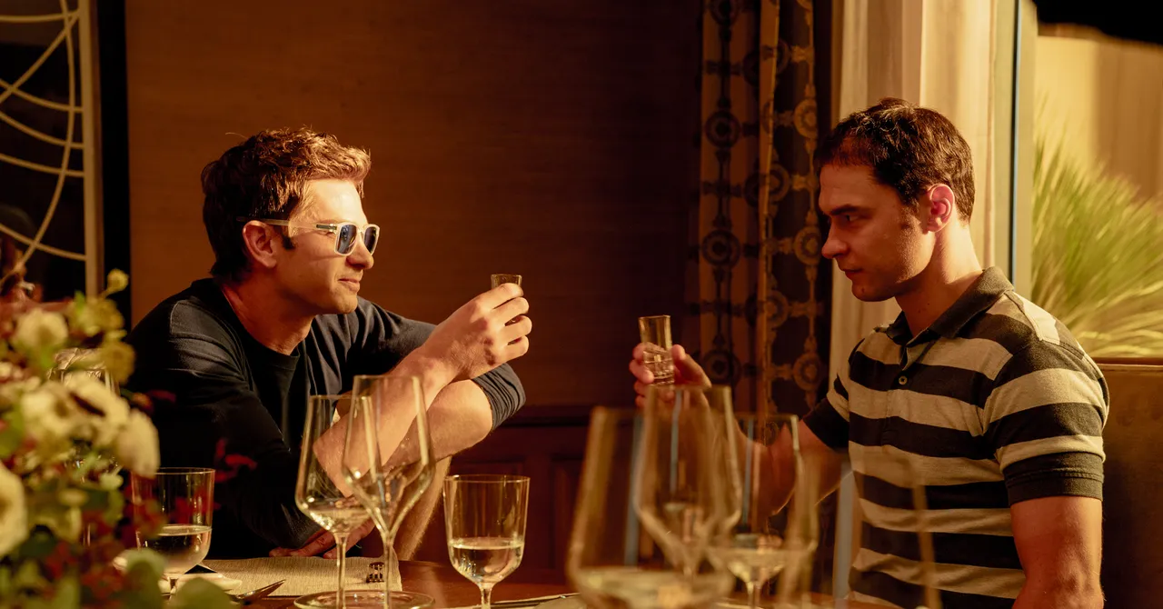 Two men seated at a table, raising glasses, surrounded by wine glasses and floral decorations in a warmly lit room.