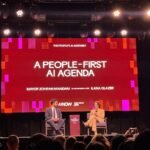 Two people seated on stage under bright lights, discussing AI policy. Screen displays "A People-First AI Agenda".
