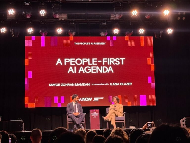 Two people seated on stage under bright lights, discussing AI policy. Screen displays "A People-First AI Agenda".