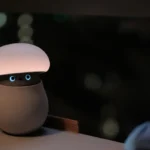 Mushroom-shaped AI robot lamp with glowing top and blue eyes on a desk, person looking at it in dimly lit room.