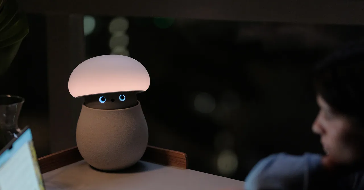 Mushroom-shaped AI robot lamp with glowing top and blue eyes on a desk, person looking at it in dimly lit room.