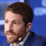 Man with a beard and a headset microphone speaks at an event; blurred text in background reads "global conference".