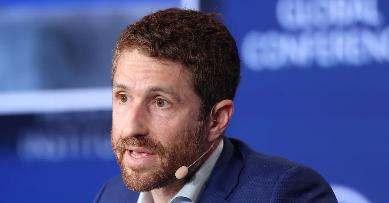 Man with a beard and a headset microphone speaks at an event; blurred text in background reads "global conference".