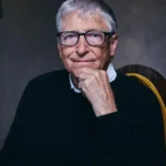 An older man with glasses and a sweater sits thoughtfully with his hand on his chin, against a neutral background.
