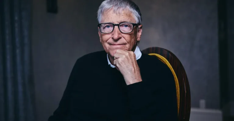 An older man with glasses and a sweater sits thoughtfully with his hand on his chin, against a neutral background.