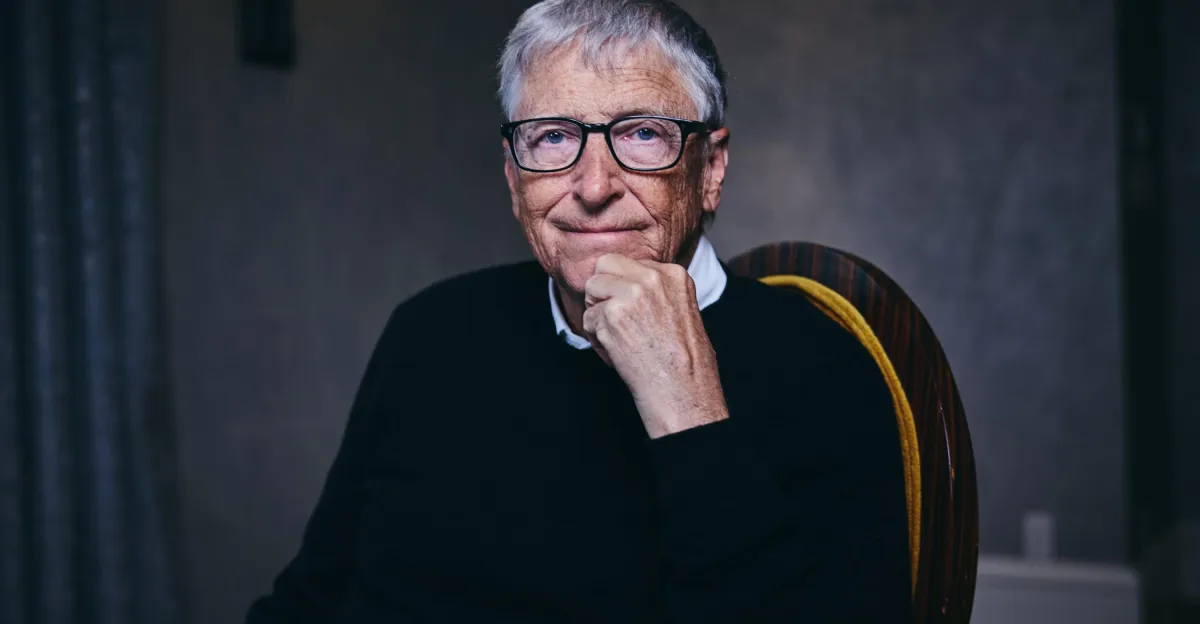 An older man with glasses and a sweater sits thoughtfully with his hand on his chin, against a neutral background.