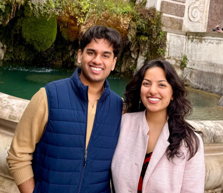 Two people smiling in front of a stone fountain with greenery and water, one holding a smartphone camera.