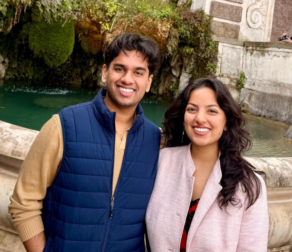Two people smiling in front of a stone fountain with greenery and water, one holding a smartphone camera.