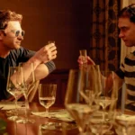 Two men seated at a dining table, toasting with small glasses. One wears sunglasses, surrounded by empty wine glasses.