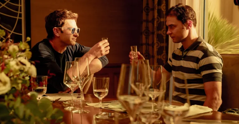 Two men seated at a dining table, toasting with small glasses. One wears sunglasses, surrounded by empty wine glasses.