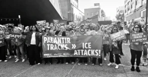 Crowd of protesters holding signs that say "Palantir Tracks >> ICE Attacks!" in a city street setting.