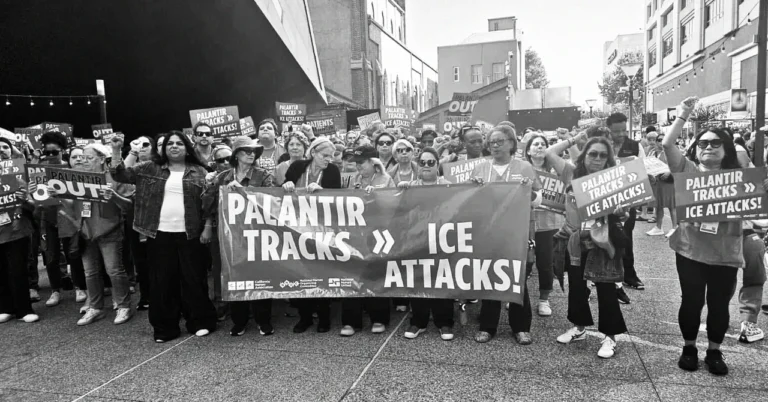 Crowd of protesters holding signs that say "Palantir Tracks >> ICE Attacks!" in a city street setting.