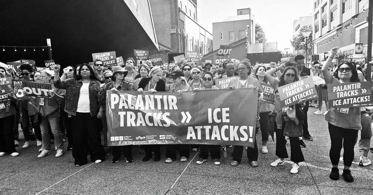 Crowd of protesters holding signs that say "Palantir Tracks >> ICE Attacks!" in a city street setting.