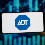 Smartphone displaying ADT logo with a digital stock market chart in the background.