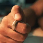 A hand wearing a smart ring with a button, resting on a table, blurred background.