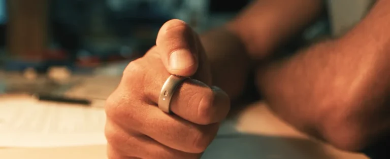 A hand wearing a smart ring with a button, resting on a table, blurred background.