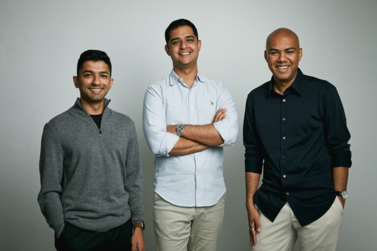 Three men standing and smiling against a plain background, wearing casual and semi-formal attire.