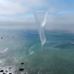 White weather balloon floating over a coastal ocean with rocky shoreline, under a cloudy blue sky.