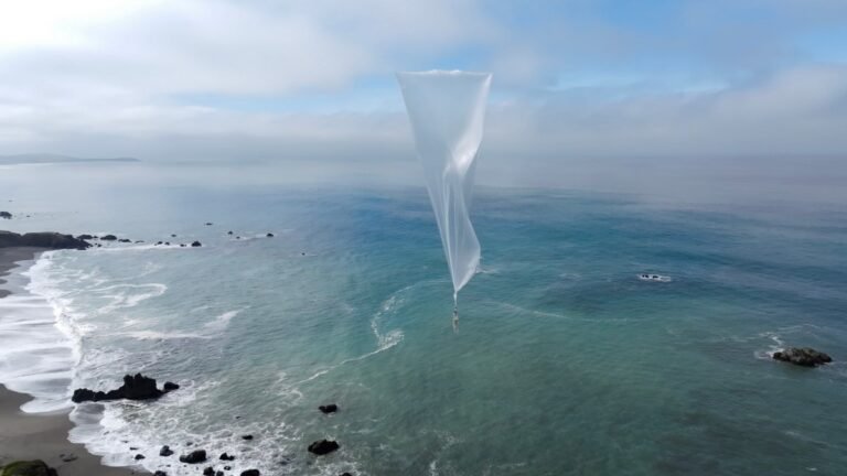 White weather balloon floating over a coastal ocean with rocky shoreline, under a cloudy blue sky.