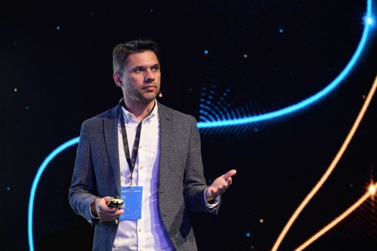 Man in a suit speaking on stage with a headset, holding a clicker, abstract light patterns in the background.