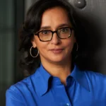 Person with glasses and hoop earrings, wearing a blue shirt, standing against a dark background.